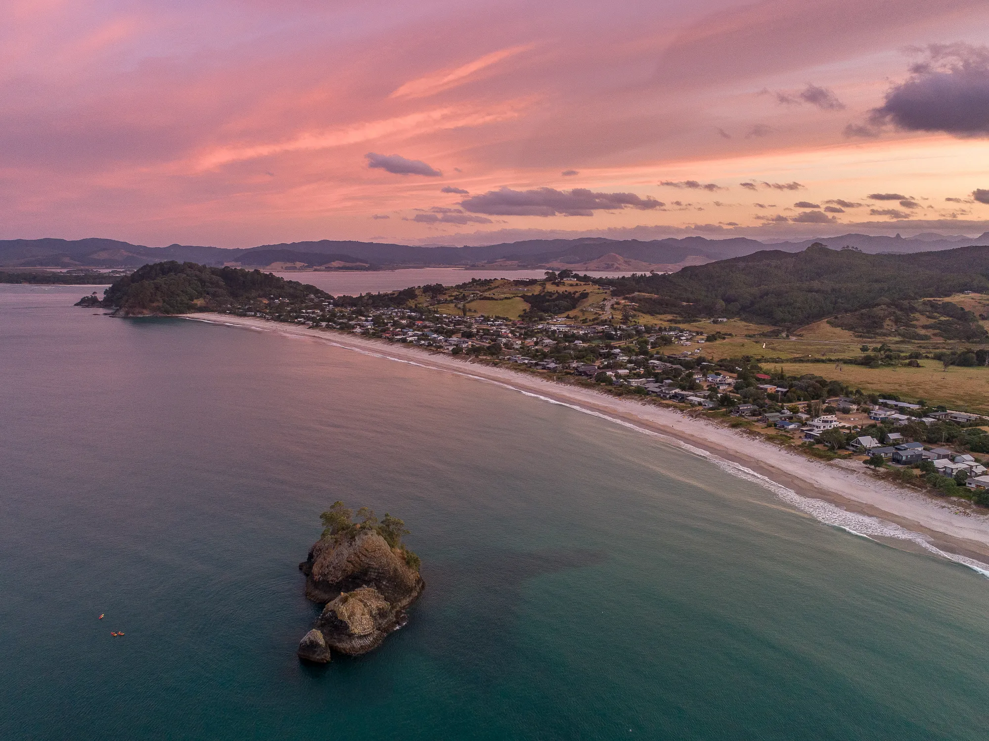 An aerial view of Whangapoua beach and village beside a rocky islet under a pink sunset